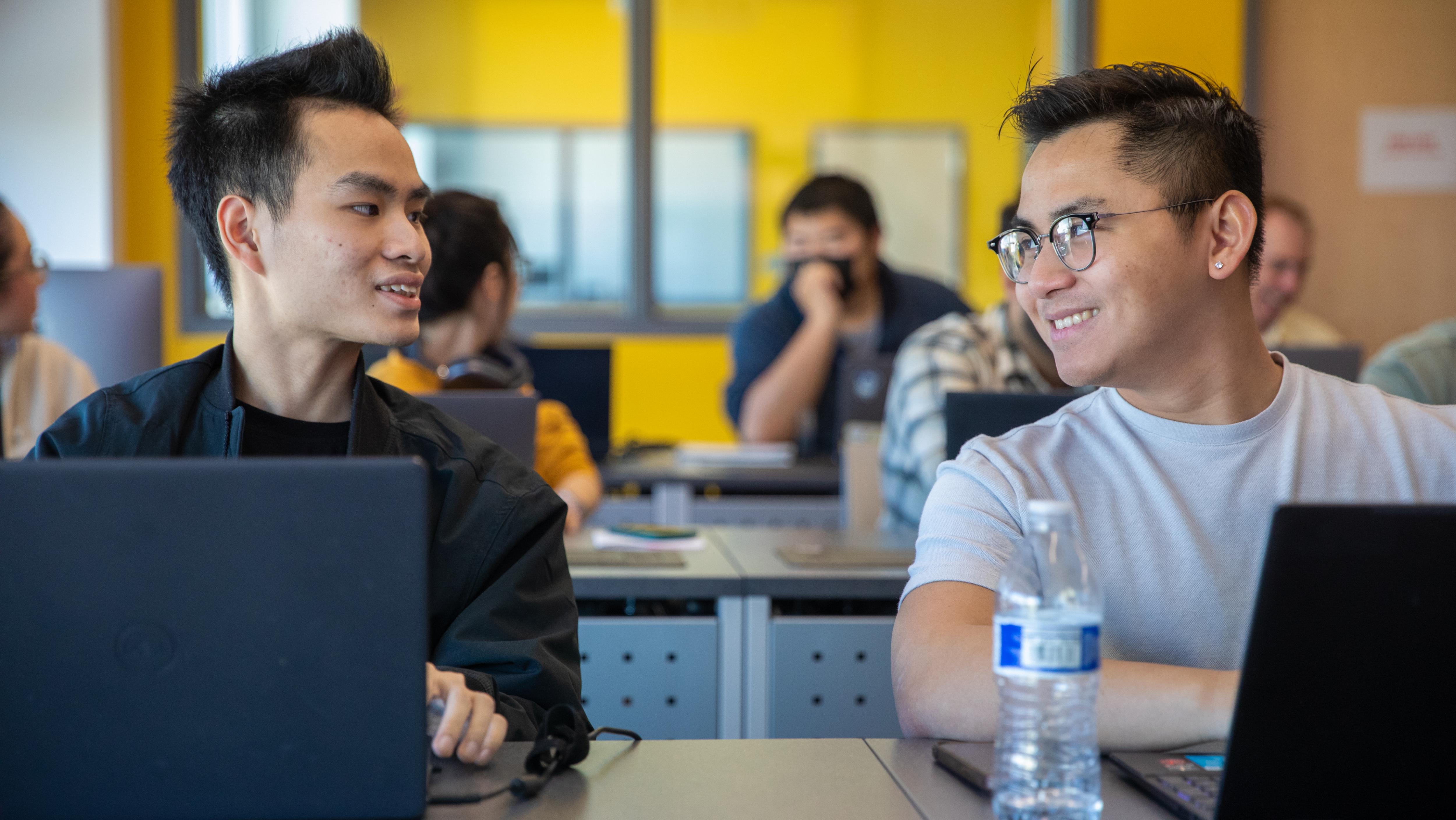 Two male students seated side-by-side at classroom desks, turning toward each other and smiling while conversing in front of their laptops.