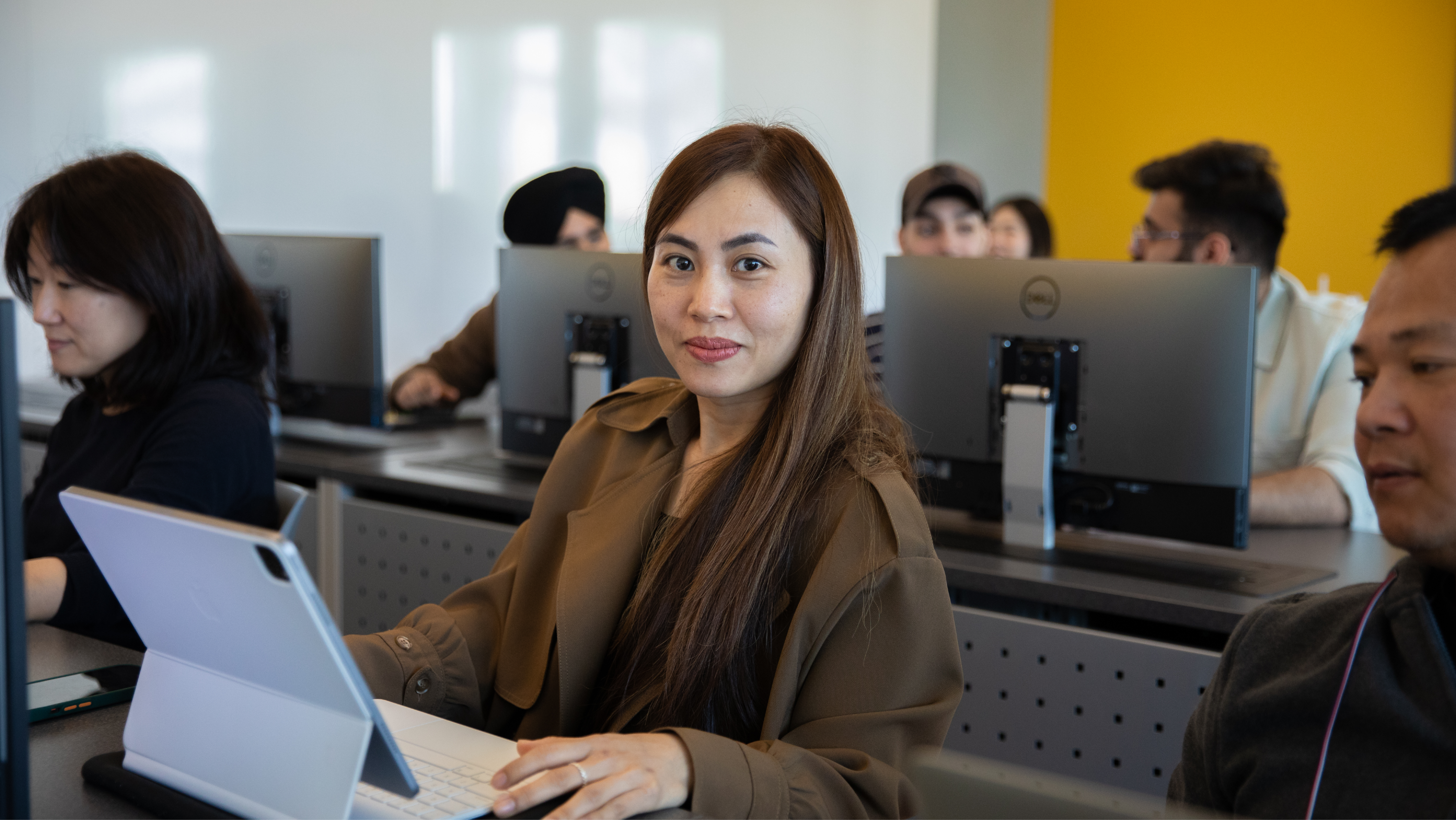A woman in a brown jacket sitting at a computer lab desk, smiling directly at the camera while using a tablet with a keyboard case.