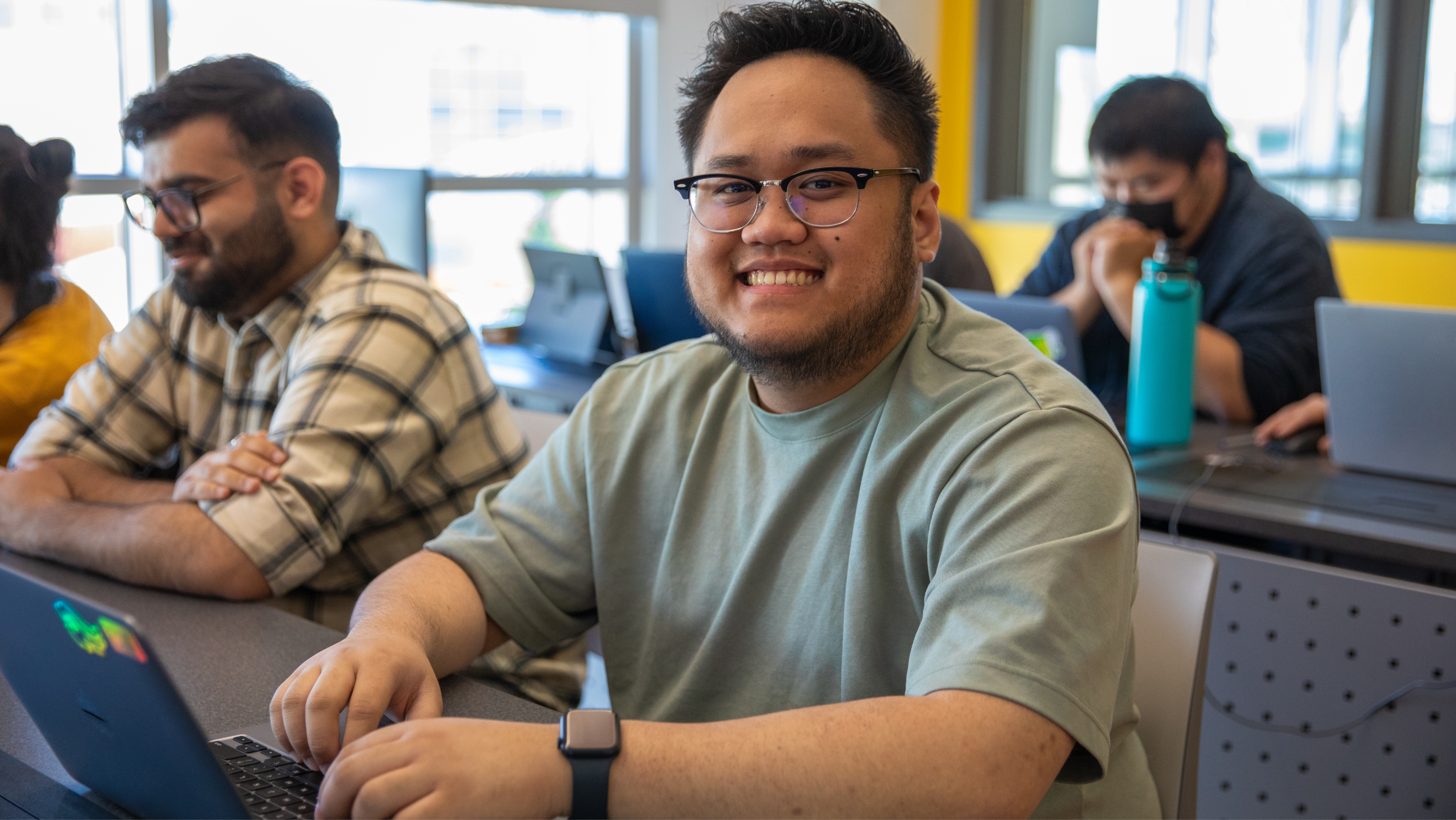 A male student wearing glasses and a light green t-shirt sitting at a desk in a classroom, smiling at the camera while typing on his laptop.