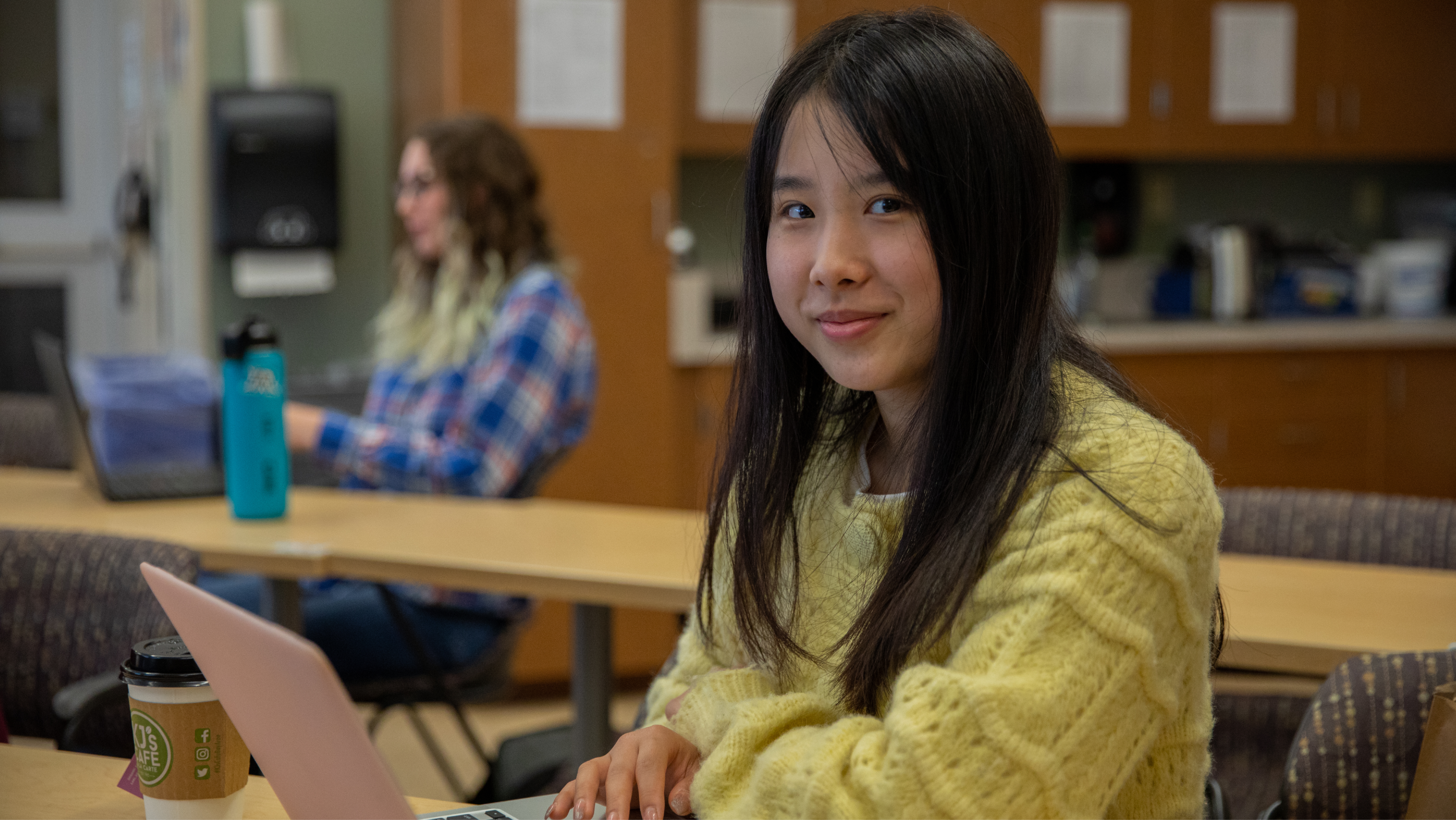 A young woman wearing a yellow knit sweater sitting at a classroom table with a coffee cup, looking towards the camera with a soft smile.