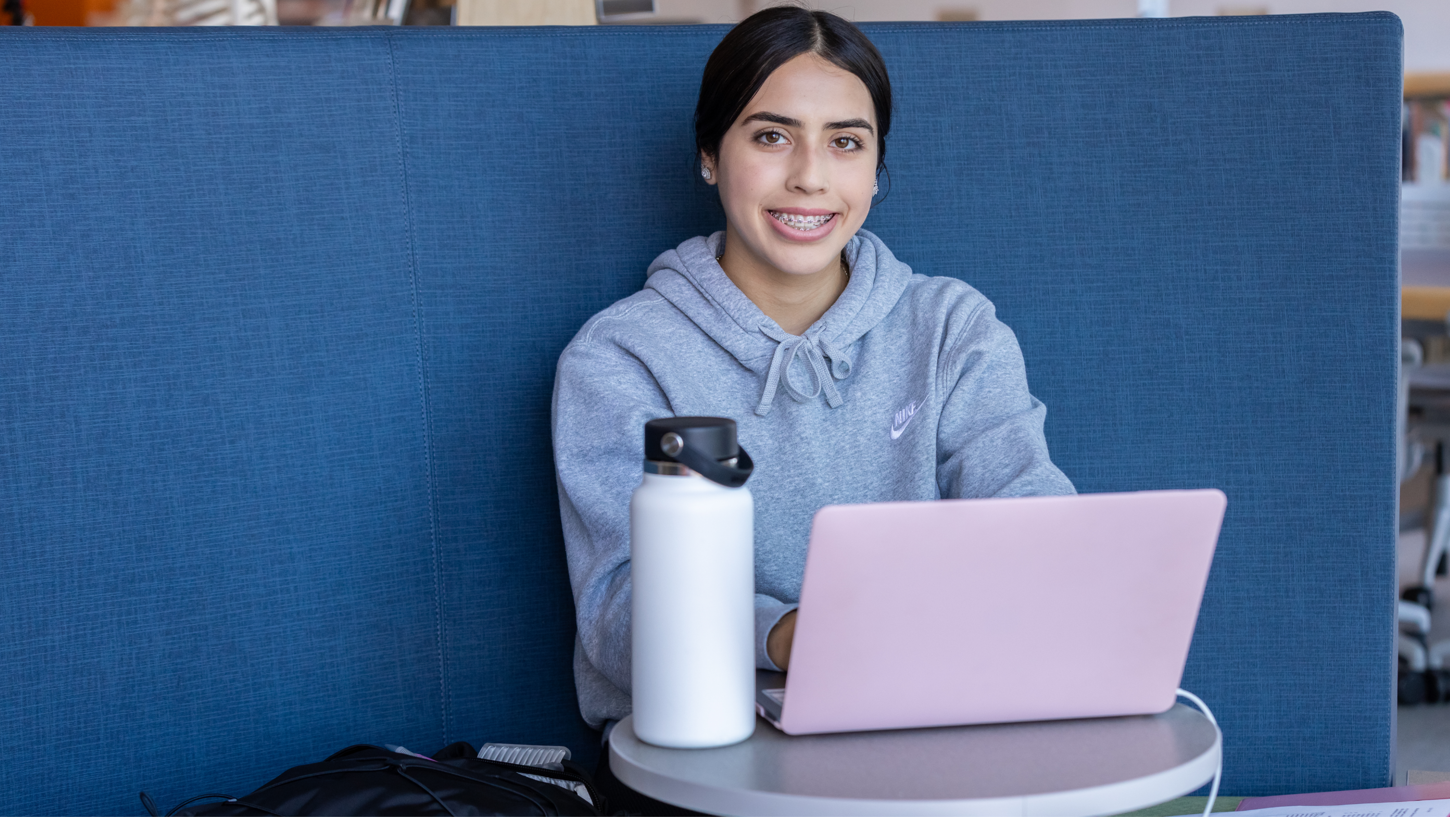 A female student sitting at a tall table near a bright window, smiling as she works on a laptop with earbuds in.