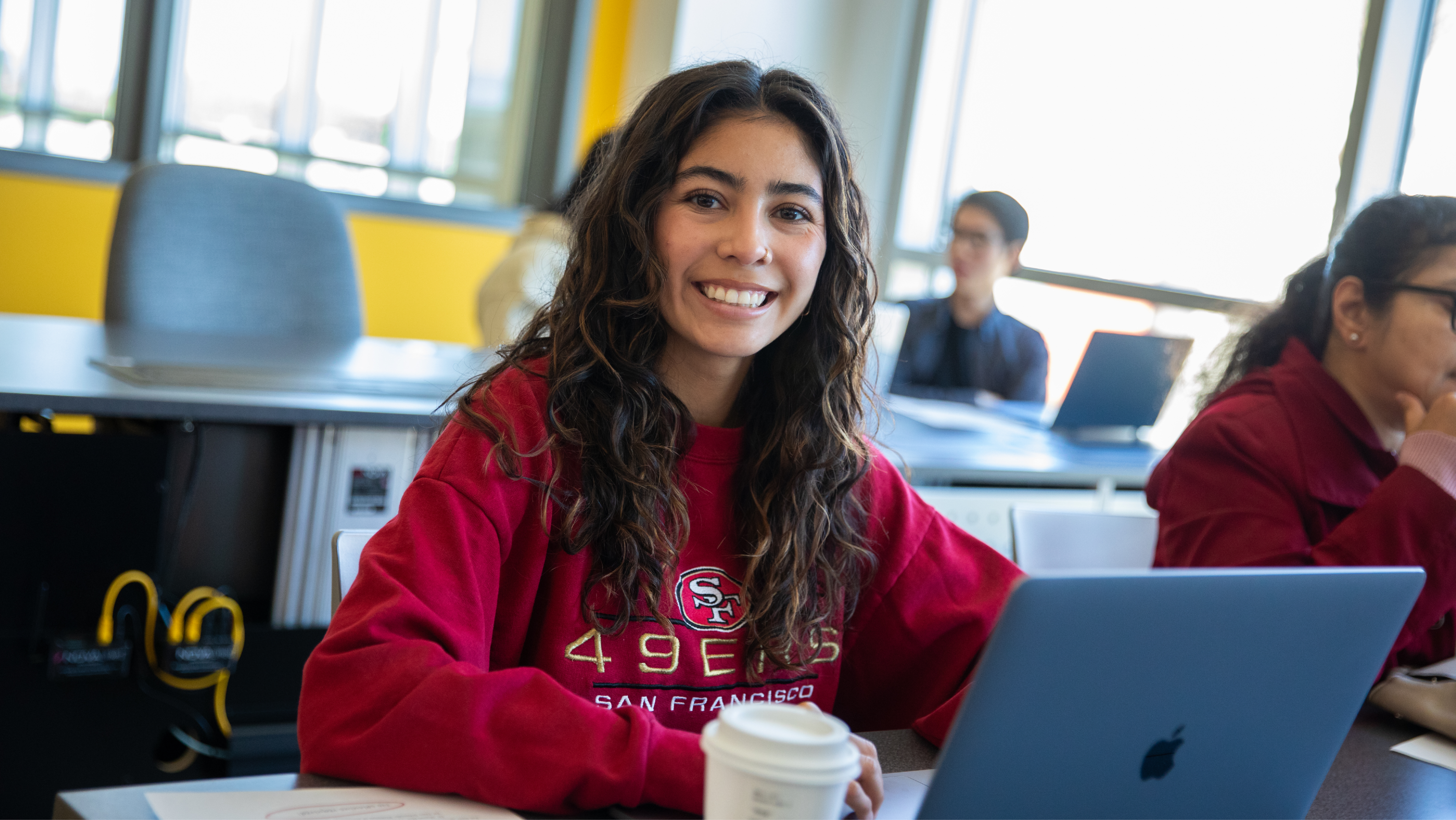 A female student with long wavy hair wearing a red 49ers sweatshirt, smiling warmly at the camera next to her open laptop and coffee cup.