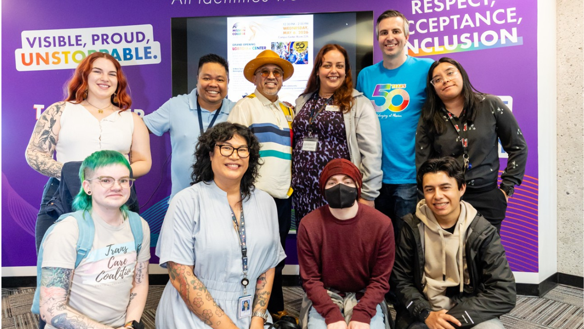 A diverse group of people gathering for a photo inside the LGBTQIA+ Center in front of a purple wall that reads "LGBTQIA+ Center - All identities welcome."