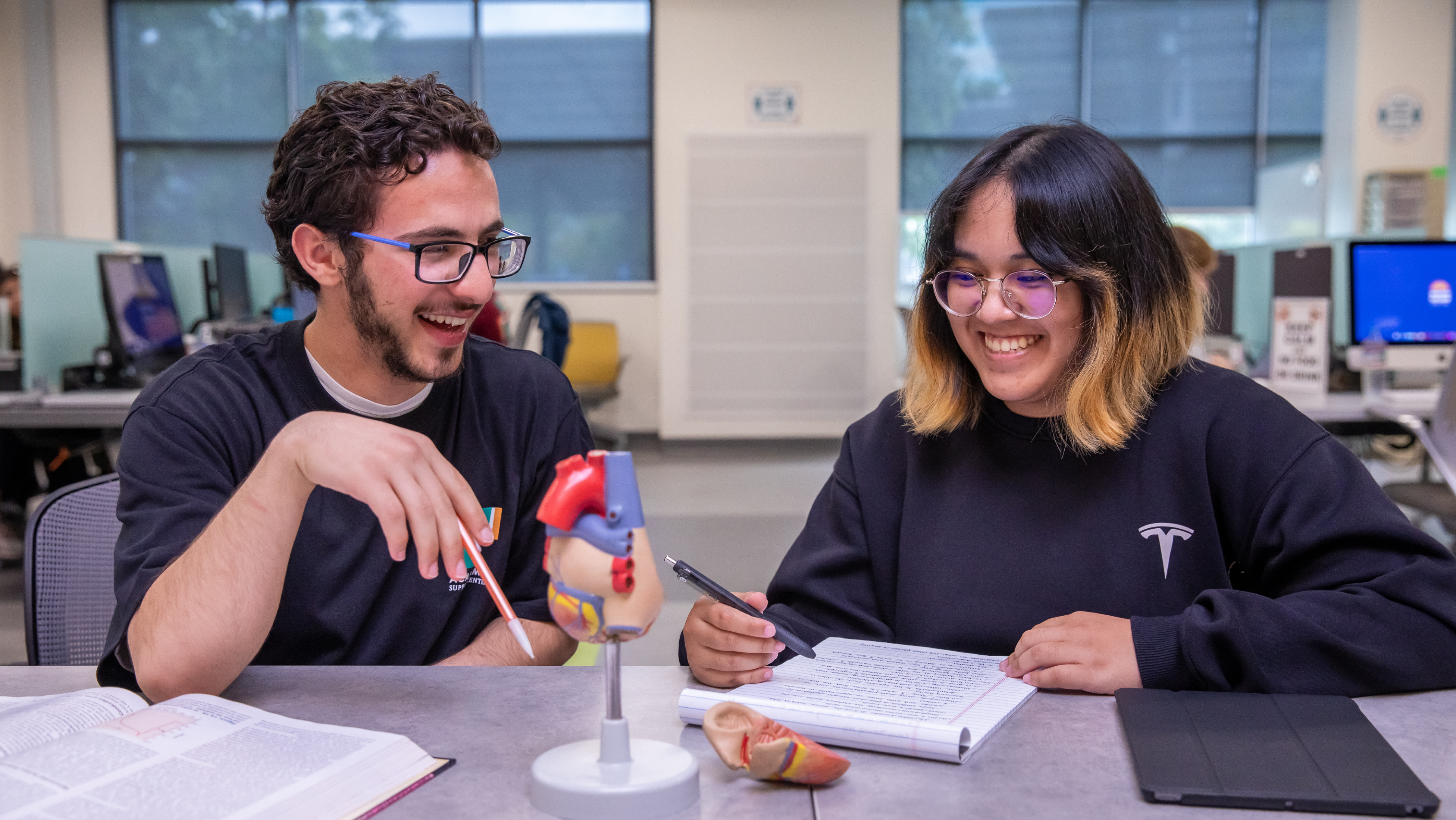 Two smiling students sitting at a table in a study center, pointing at an anatomical heart model while working together with open textbooks and a notebook.