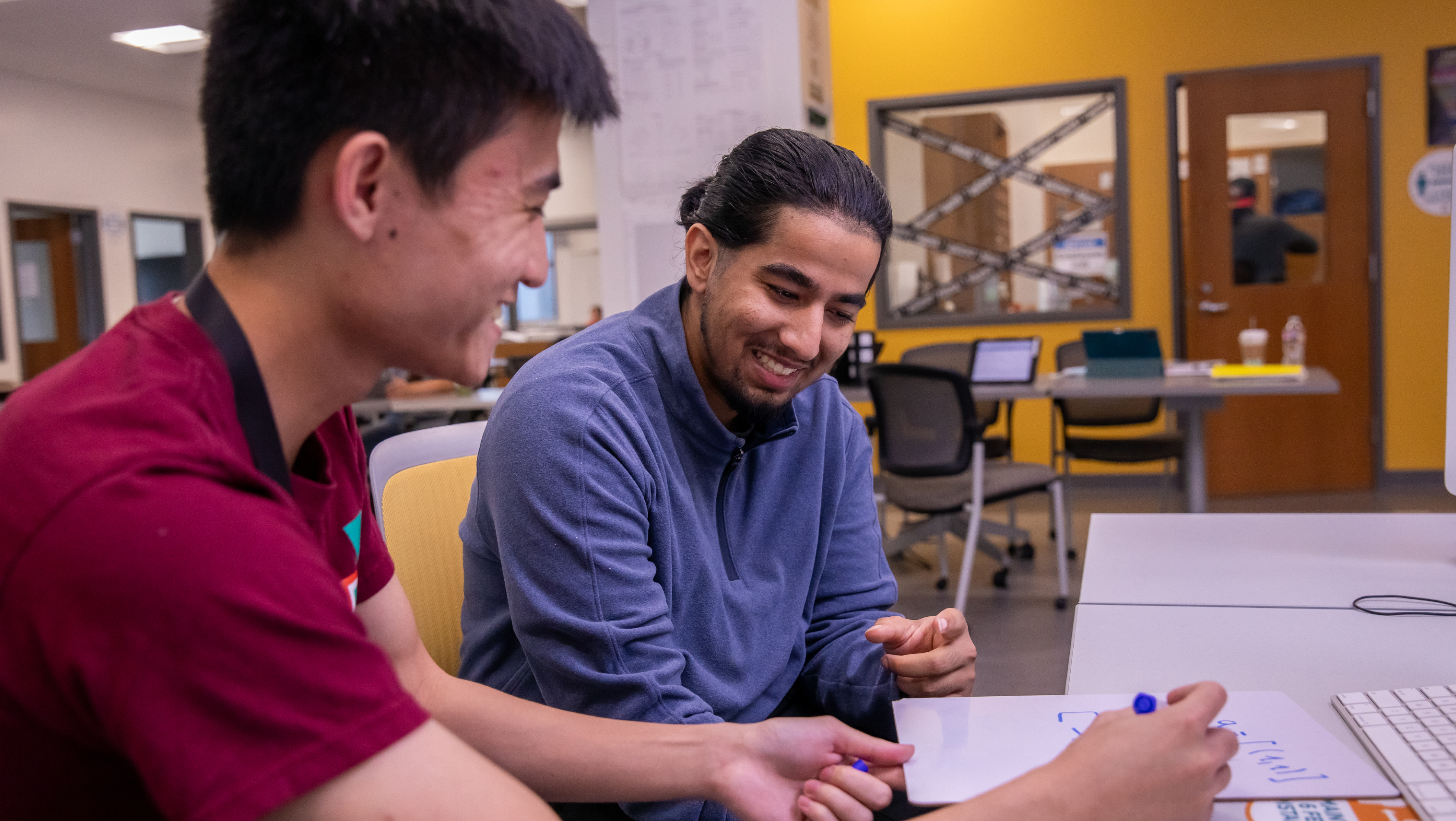 Two male students working collaboratively at a desk, smiling as one writes out a formula on a small dry-erase board.