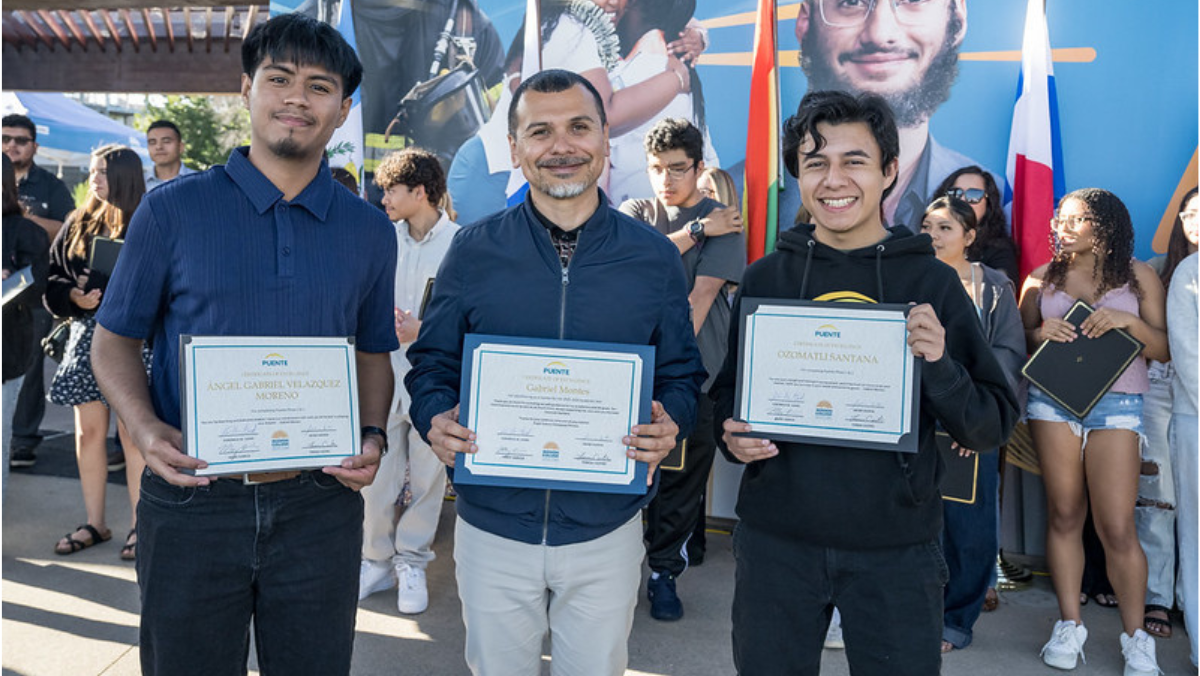 Three men standing side by side outdoors holding framed Puente certificates of achievement with various national flags displayed behind them.