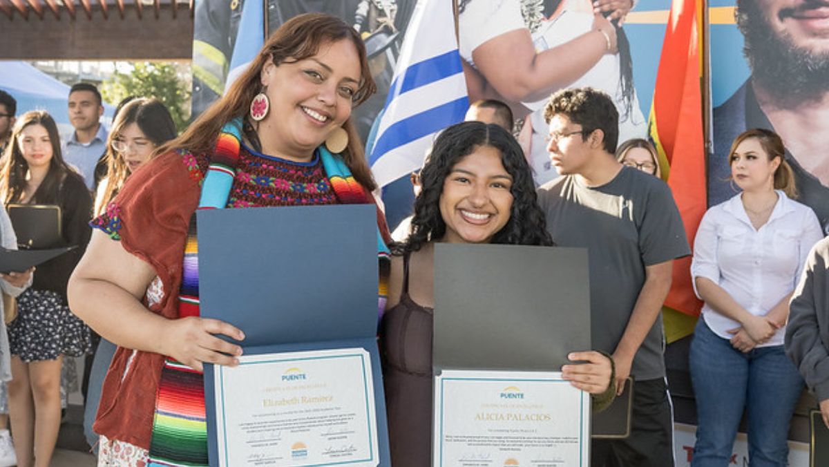 Two women smiling while holding framed Puente Program certificates of achievement at an outdoor event.