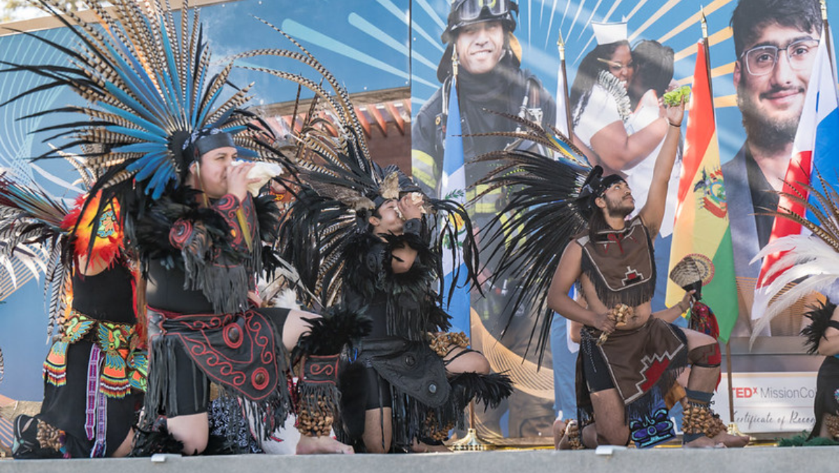 Aztec dancers in traditional regalia, feather headdresses, and shell instruments performing on an outdoor stage.