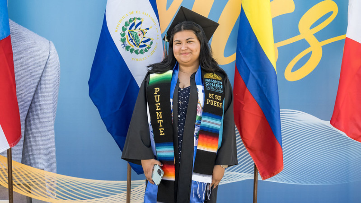 A smiling female graduate wearing a black cap and gown with a colorful "Puente" graduation stole, standing in front of international flags.