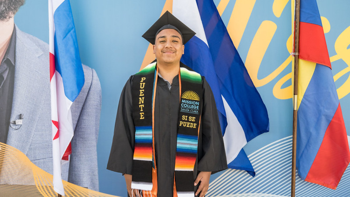 A smiling male graduate wearing a black cap, gown, and "Puente / Sí Se Puede" graduation stole, standing in front of national flags.