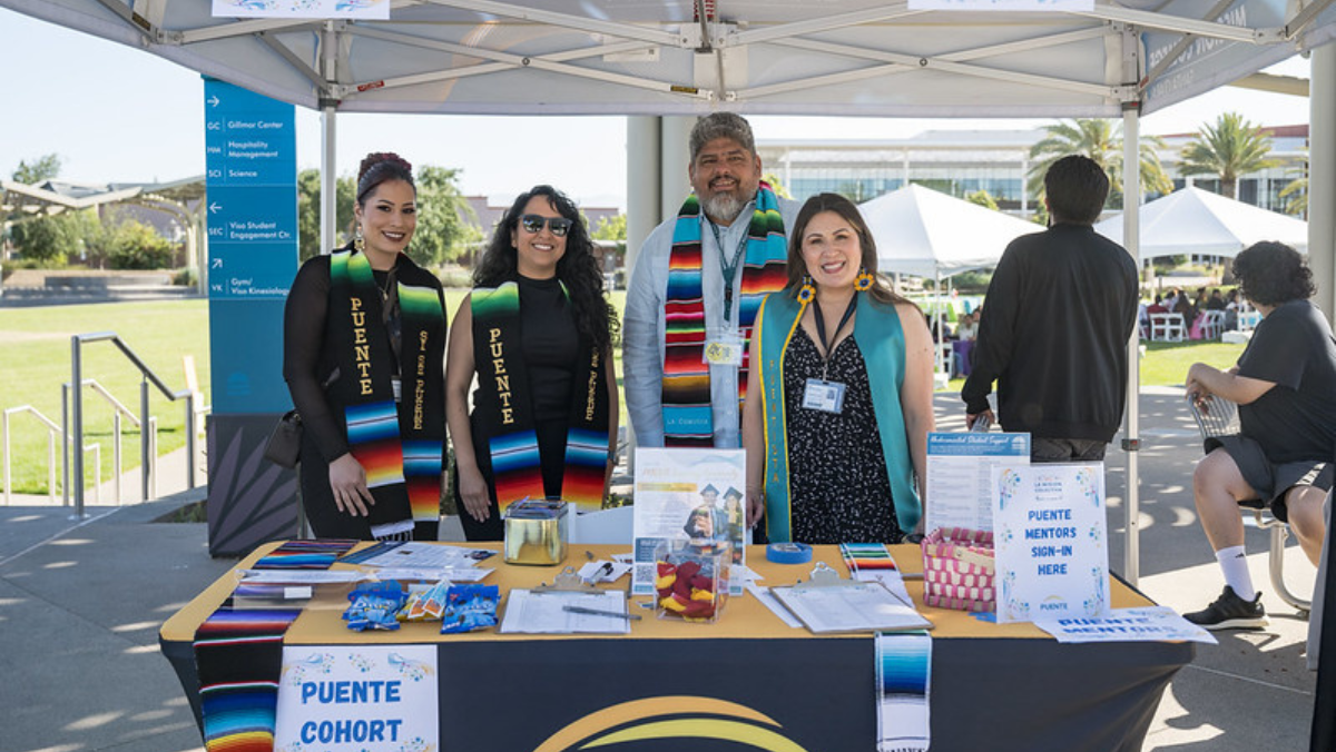Four staff members and mentors wearing colorful serape stoles standing behind a check-in table outdoors with a "Puente Cohort" banner and sign-in sheets.