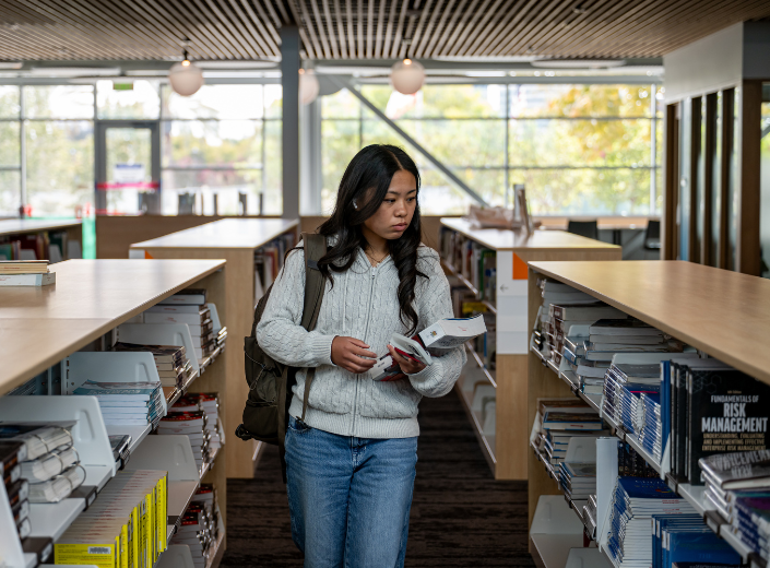 A college student with a backpack carries books while walking between modern wooden bookshelves inside the naturally lit Mission College Library.